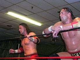 Two white males wearing red wrestling gear standing in a wrestling ring with red and black ropes.
