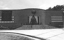 A black and white picture of a one story brick building. The entrance door is in the center of the building and three rows of white diamond shaped tiles decorate either side.