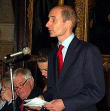A balding man in his 40s, with close-cut brown hair, wearing a suit with a red tie, speaks into a microphone with papers in his hands