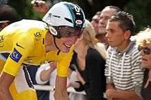 A road racing cyclist in a yellow skinsuit, wearing an aerodynamic helmet. His bicycle is not visible. Spectators watch from the roadside.