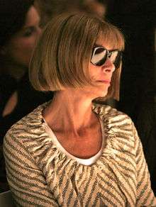Anna Wintour wearing sunglasses and a grey-and-white striped top in a dark background looking to the right