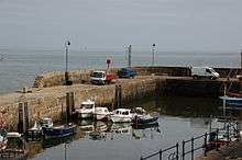 Small, stone-walled harbour containing a handful of small pleasure and fishing boats, with the open sea pictured beyond.