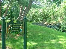 photograph of a multi-coloured carved wooden sign which reads "Anne Hutchinson/Mary Dyer Memorial Herb Garden," behind which is a scenic small waterfall surrounded by green foliage.