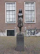 A bronze statue of a smiling Anne Frank, wearing a short dress and standing with her arms behind her back, sits upon a stone plinth with a plaque reading "Anne Frank 1929–1945". The statue is in a small square, and behind it is a brick building with two large windows, and a bicycle. The statue stands between the two windows.