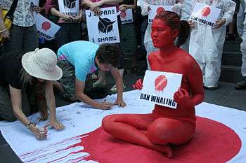 An oriental woman has painted herself red holding a sign (while sitting down) that says "Ban Whaling" while a crowd around her signs a petition. She is sitting on a Japanese flag with red dripping down (presumably to symbolize blood)