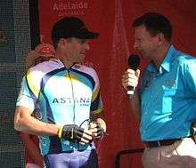 A man in his late thirties in a blue and white cycling jersey with yellow trim stands next to a man in his mid-fifties wearing a light blue collared shirt and holding a microphone. They stand in front of a red backdrop, and a woman in a cowboy hat is partly visible behind the cyclist.