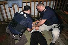 Two police officers seen from above and behind, on their knees putting handcuffs on a prone man between them.