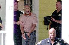 On the porch of a yellow-clapboard house stands a middle-aged African American man in polo shirt and slacks, in the middle of a statement, his mouth agape, his hands handcuffed to the front. Behind him stands a uniformed Caucasian policeman, lightly grasping the man's upper arm. Facing the pair is an officer to the right, his left hand held up in a "hold on" gesture. Another African American officer is in the foreground, below the level of the porch, with his arms akimbo and his back to the scene while he faces the general direction of the camera.