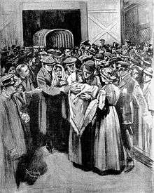 A man wearing a bowler hat and a woman in a shawl embrace among a crowd of people standing in a wooden building