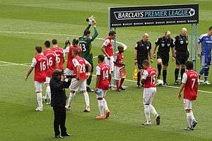 A coloured photograph of a group of men, dressed in red. They are walking in order to form a line. A man dressed in a green shirt places his hands together and raises them, in direction to his right. To the far right of the photograph, there is a man dressed in a blue kit, in line with three men who are dressed in black. Behind them is a placard with the words 'Barclays Premier League'.