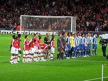 Two football teams and a refereeing team lining up in a football stadium pitch. The leftmost team wears red shirts and white shorts and socks, while the other wears blue shorts and socks and shirts with vertical blue-and-white stripes. The refereeing trio wears light gray equipment. A goal and a packed stand behind it are seen in the background.