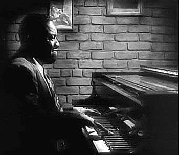 A short-haired black man is sitting behind a piano facing right. He is wearing an opened suit jacket, a white shirt and a necktie. His hands are on the keyboard and he appears to be playing. On the background there is a brick wall on which two paintings or photographs are partly visible.