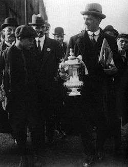 A tall man wearing a hat, shirt and tie and overcoat, holding a trophy. Next to and behind him are a crowd of people, looking in his direction.
