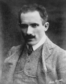 A head-and-shoulders photograph of a man in a three-piece suit with tie and watch-chain. His intense-looking appearance is complemented by receding dark curly hair and a curled moustache.