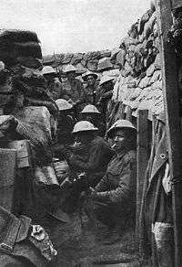 Black and white photo of a group of men wearing military uniform, including helmets, in a trench. Four men are crouching on the floor of the trench and another four are standing.