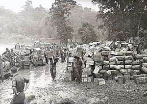 Black and white photo of a large number of small wooden crates stacked on muddy ground, with men wearing military uniforms carrying additional small wooden crates.