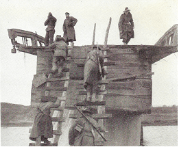 Three soldiers are standing on top of a partially destroyed bridge, while other soldiers climb up two roughly constructed ladders with their rifles slung over their shoulders while carrying stores.