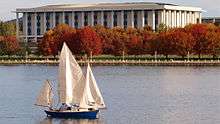 The national library is a rectangular building with tall pillars similar to a Roman/Greek style building. It stands on the shores of a landscaped lake surrounded by deciduous trees with red leaves.