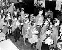  A group of small children and their teachers in a line, wearing brown capes and carrying food in their hands