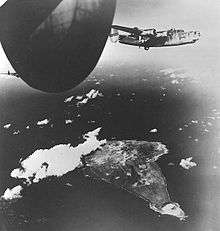 A four engined World War II-era aircraft flying above an island which is party covered in cloud as well as smoke. Sections of two other aircraft are visible.