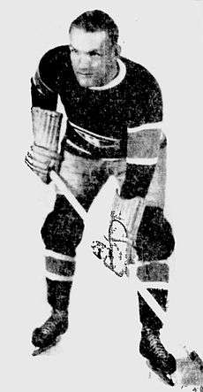 A man with short hair poses in a full ice hockey uniform, including stick and skates. He is looking slightly to his right with a serious look on his face.