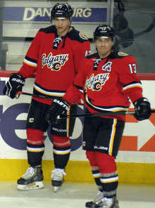Two hockey players in full uniform stand beside each other.  They are in matching red uniforms with black pants and black, white and yellow trim.  The jersey front says "Calgary" in script with a small stylized "C" logo.