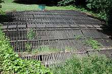 A horizontal wooden rack roughly 30 feet (9.1 m) wide and 30 feet (9.1 m) long lies in flat, grassy area.