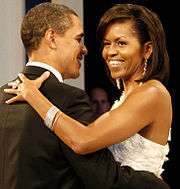 Barack and Michelle Obama dancing arm-in-arm and smiling. She is wearing a white dress, large ring, long earrings and a bracelet. He is wearing a black tuxedo.