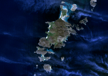 A large, dark green island seen from above is surrounded by smaller islands in a dark blue body of water. Wispy clouds partially obscure the view.