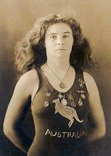 A sepia-toned photograph taken from the waist up, showing a white woman of medium build with shoulder-length wavy hair, wearing a one-piece swimming costume with the word "AUSTRALIA" on the front and a depiction of a kangaroo. She has nine medals pinned to costume around the upper chest area, and is wearing another medal around her neck.