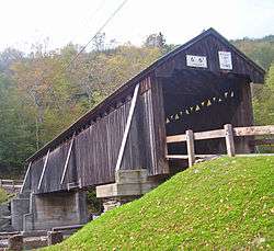 A dark brown covered bridge viewed from below and to the left of one of its portals with weight and height limitation signs
