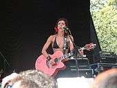 A man on a stage, wearing a black bikini top and skirt. She is standing behind a microphone stand and holding an acoustic guitar.
