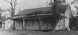 A black and white image of the meetinghouse as it originally appeared, with a long, moderately sloped roof and porch running its full length. There are less trees around it than the above image