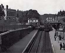 A black-and-white photograph taken probably from the station footbridge, looking down the double-track line. Half a dozen people are on the right-hand platform, the left-hand one is deserted.