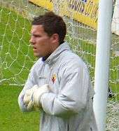 A man in a grey football shirt and goalkeeping gloves standing on a playing pitch in front of a goal.