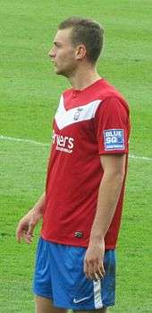 A man with brown hair who is wearing a red top, blue shorts and white socks. He is standing on a grass field.