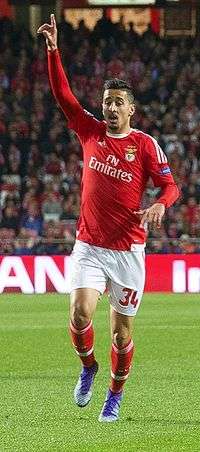 A man wearing red shirt and white shorts, raising his left arm.