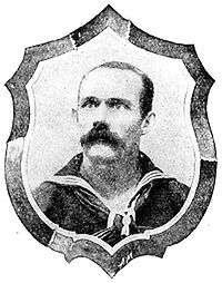 Head of a man with close cropped hair and a bushy mustache wearing a sailor suit with a wide flat collar and a scarf tied around the neck. A shield-shaped frame is drawn around the man's portrait.