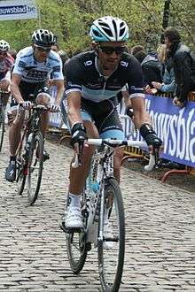 A road racing cyclist in a jersey with black sleeves, a white torso, and a blue stripe separating the two. His bicycle is only partly visible, and spectators watch from behind a roadside barricade.