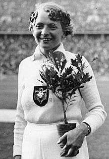 A black and white photograph of a female athlete in an all-white outfit with the Nazi eagle and swastika in the middle of her chest. She holds a small plant and wears an laurel wreath on her head.