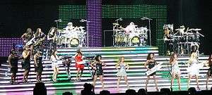A musical group of women, composed of at least eighteen of them, is performing a song on stage. They play drum kits, keyboards and saxophones, while other females sing and dance.