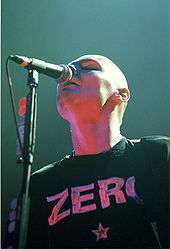 A man with a shaved head, singing into a microphone with his eyes closed. He is wearing a black shirt with the text "Zero" across the front.