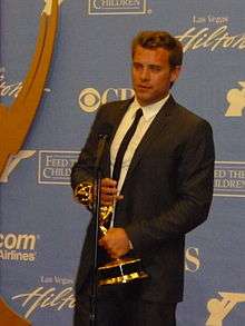 A man with dark hair, wearing a  black suit, including a black tie and white T-shirt also holding a gold statuette.
