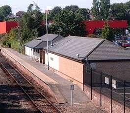 In the foreground is a single storey building with a pitched roof. A railway platform runs along the length of the building. In the background is a large red warehouse style building.