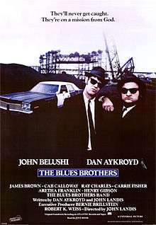 Movie poster with two of the main characters on the right-side of the image: They are both wearing black suits, hats, and sunglasses and facing forward. The man on the right is resting his arm on the shoulder of the man on the left. A police car is present on the left side of the image behind them. At the top of the image is the tagline, "They'll never get caught. They're on a mission from God." At the bottom of the poster is the title of the film, cast names, and production credits.