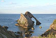A large brown rock with a striking shape lies just beyond a rock coast in a blue sea under a pale blue sky. The left hand and foreground part of the rock is wedge shaped and the sedimentary rocks it is made off are set at a 45 degree angle to the horizontal. A second part of the rock is in the shape of an arch with a thick top section and a thin downward leg. The whole structure has a strangely contrived air, suggestive of a wrecked ship.
