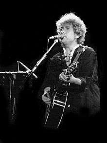 Black and white image of a man with curly hair playing an acoustic guitar and standing behind a microphone stand