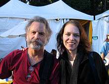 A bearded man who looks to be in his 60s wearing a dark red shirt and Welch, smiling, with her arm around him. Huge white tents are in the background.