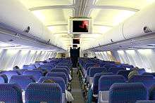 Airliner cabin. Rows of seats arranged between a center aisle.  There are overhead monitors.