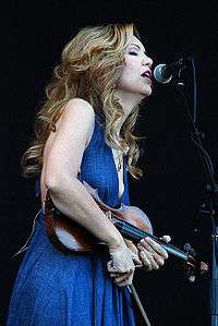 A woman in a blue dress holding a fiddle sings into a microphone